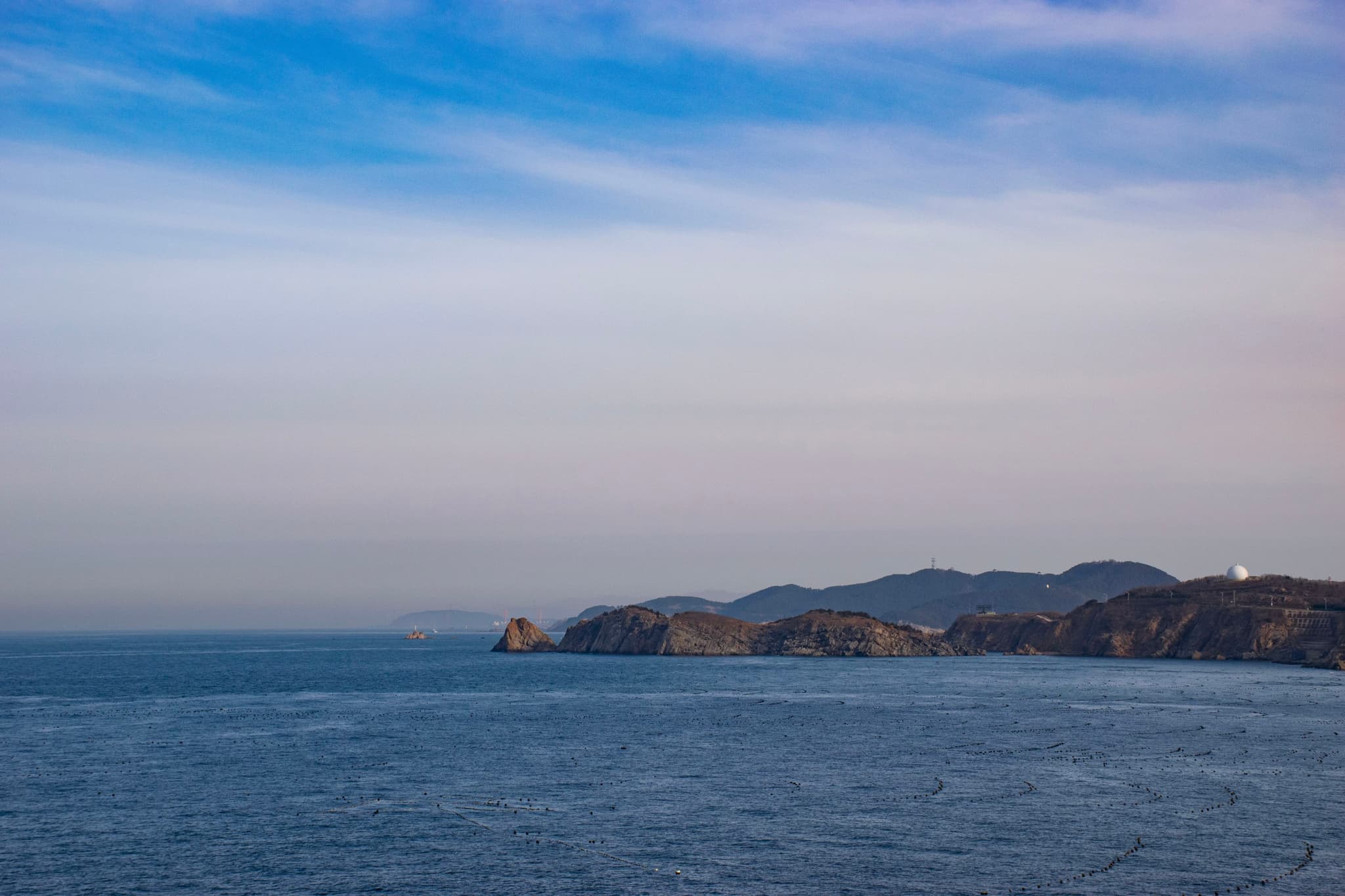 海面平静，远处的山峦在天边若隐若现，天空晴朗，微带云彩。