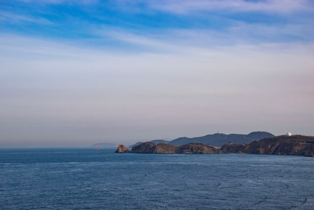 海面平静，远处的山峦在天边若隐若现，天空晴朗，微带云彩。