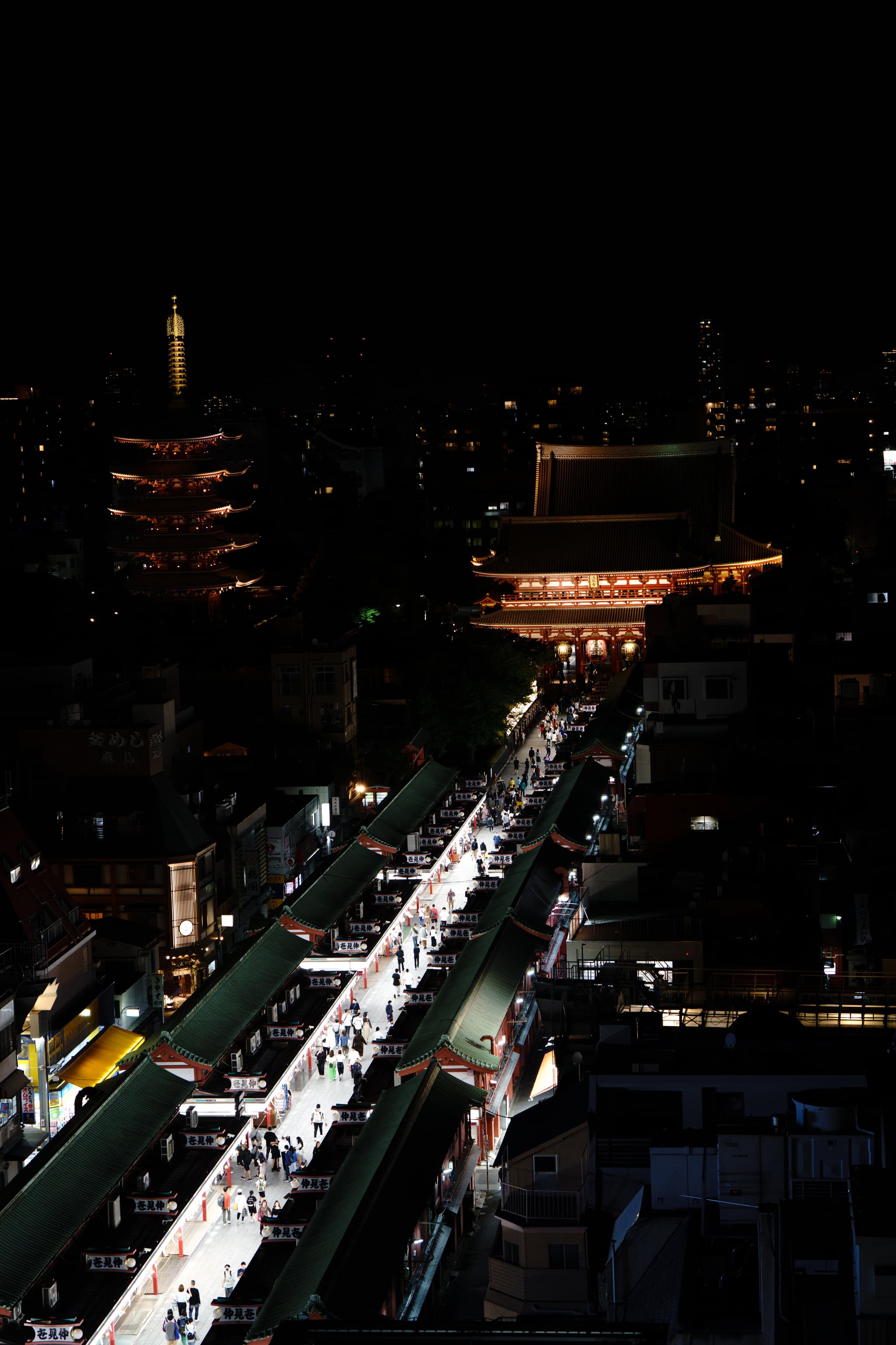 夜晚的城市景观，灯火通明的街道延伸至远处，背景中有一座传统建筑和一座塔楼。