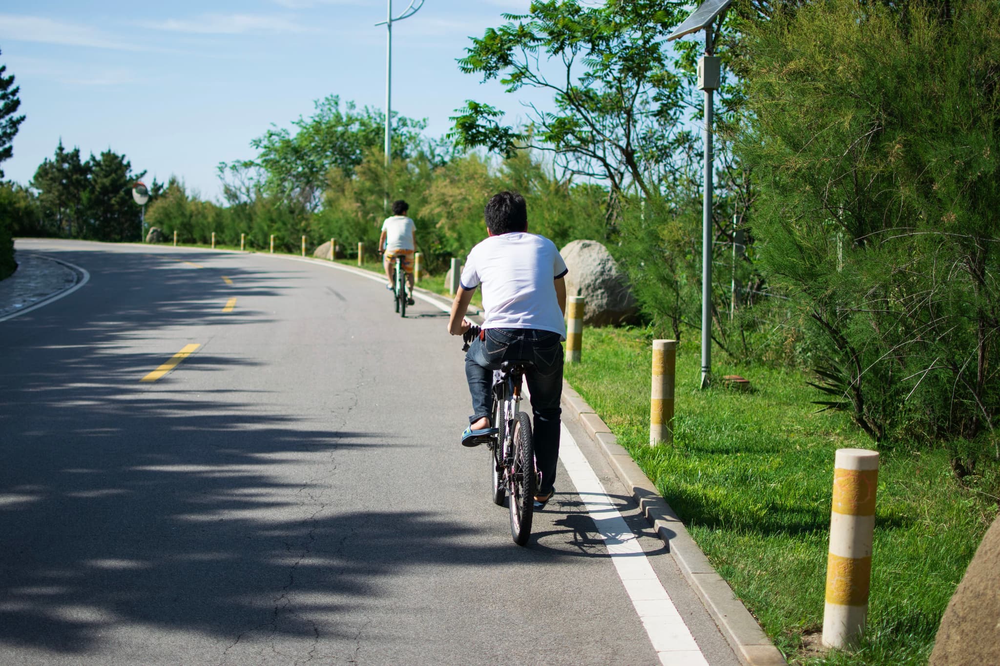 Two cyclists ride on a paved road surrounded by greenery, with one cyclist in the foreground and another further ahead