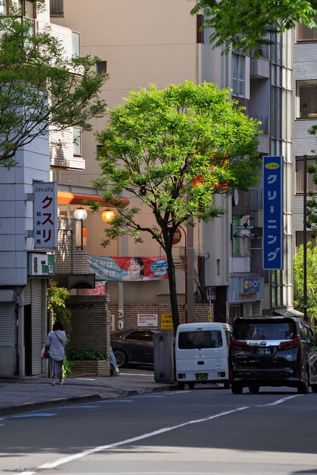 城市街道场景，阳光明媚，路边停着几辆车，行人走在路旁。街道两侧是现代建筑，树木郁郁葱葱。广告牌上有日文标识。