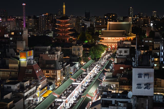 夜晚的城市景象，灯火通明，街道上人来人往，远处有高塔和寺庙建筑。