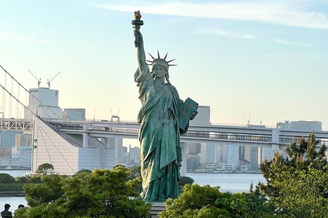 自由女神像矗立在前景，背景是城市天际线和桥梁，周围有绿色植被。