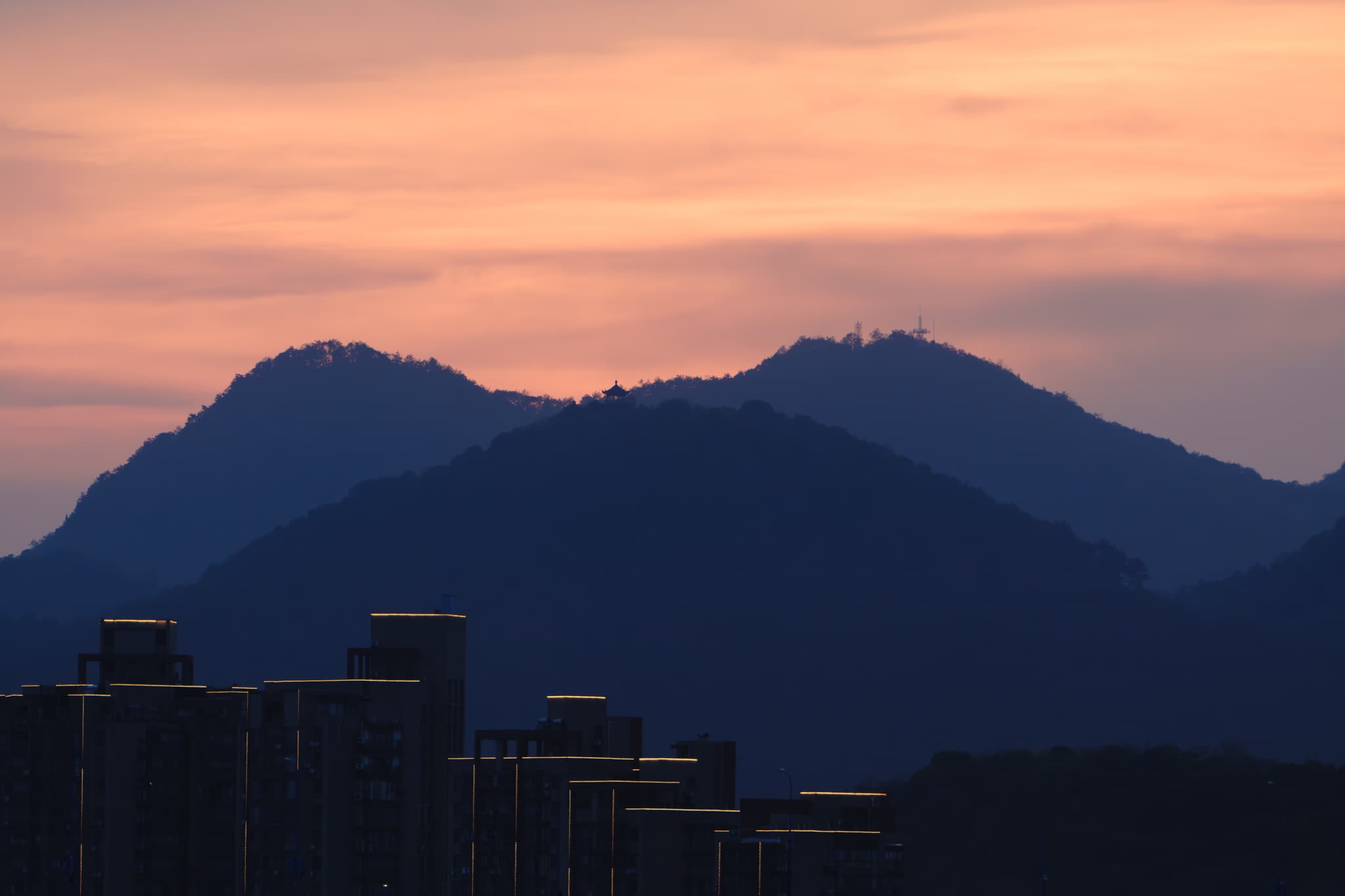 夕阳下的山峦剪影，背景是橙色和粉色的天空，前景有几栋高楼。