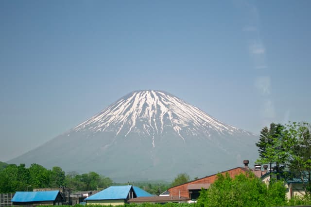 A snow-capped mountain stands tall in the background, with a few buildings and trees in the foreground under a clear blue sky