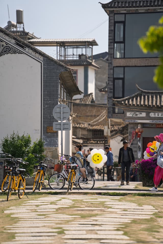 古色古香的建筑群，街道上停放着共享单车，行人手持大大的笑脸气球。