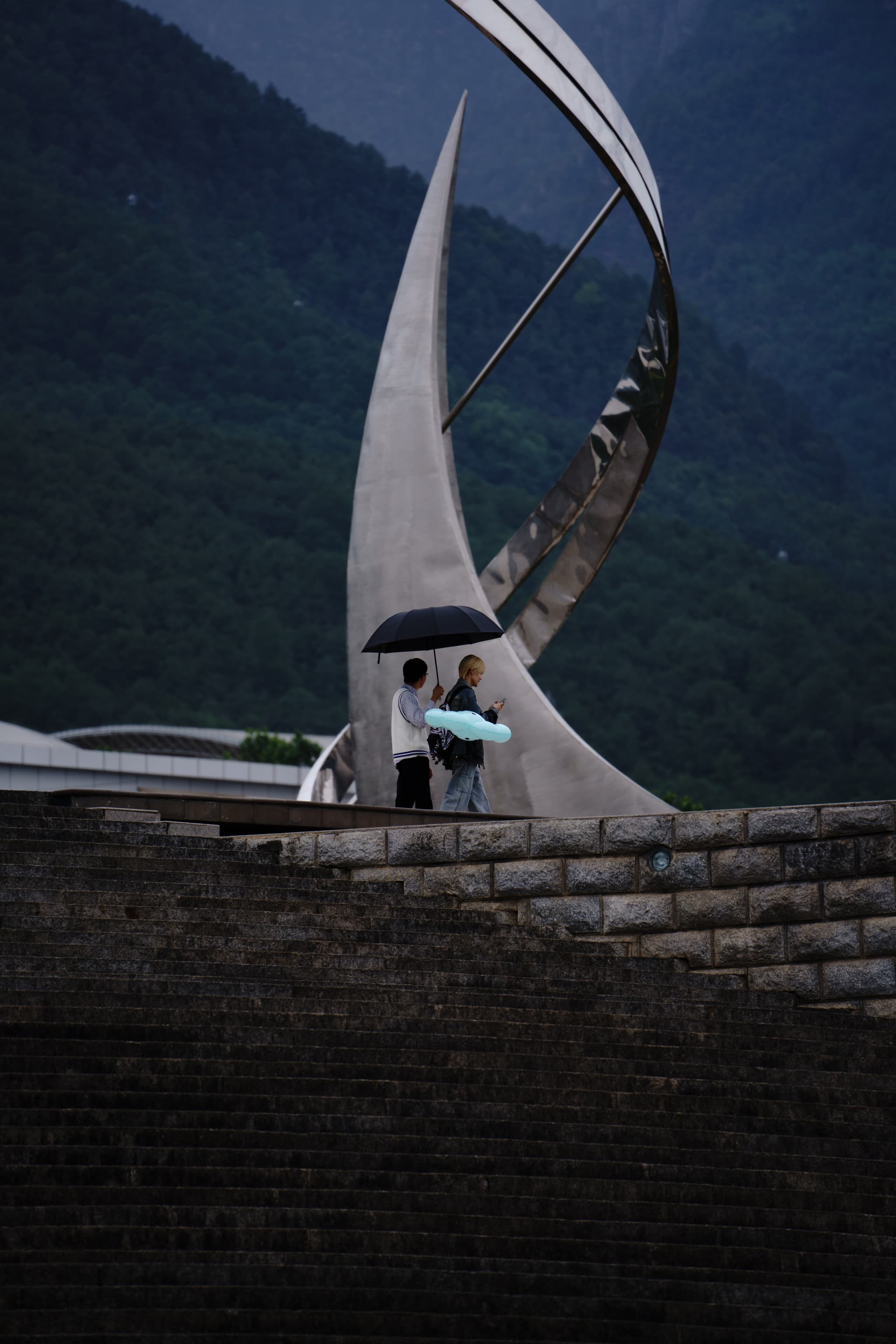 一对情侣在雨中撑伞站在现代雕塑前，背景是青山。