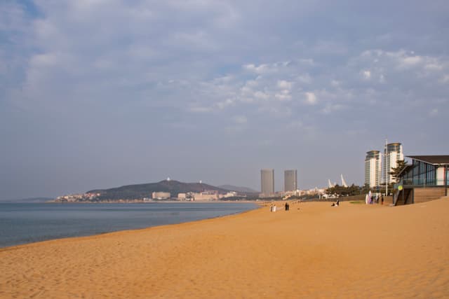 海滩上沙子金黄，远处有几栋高楼和一座小山，海水平静，天空有少量云朵。