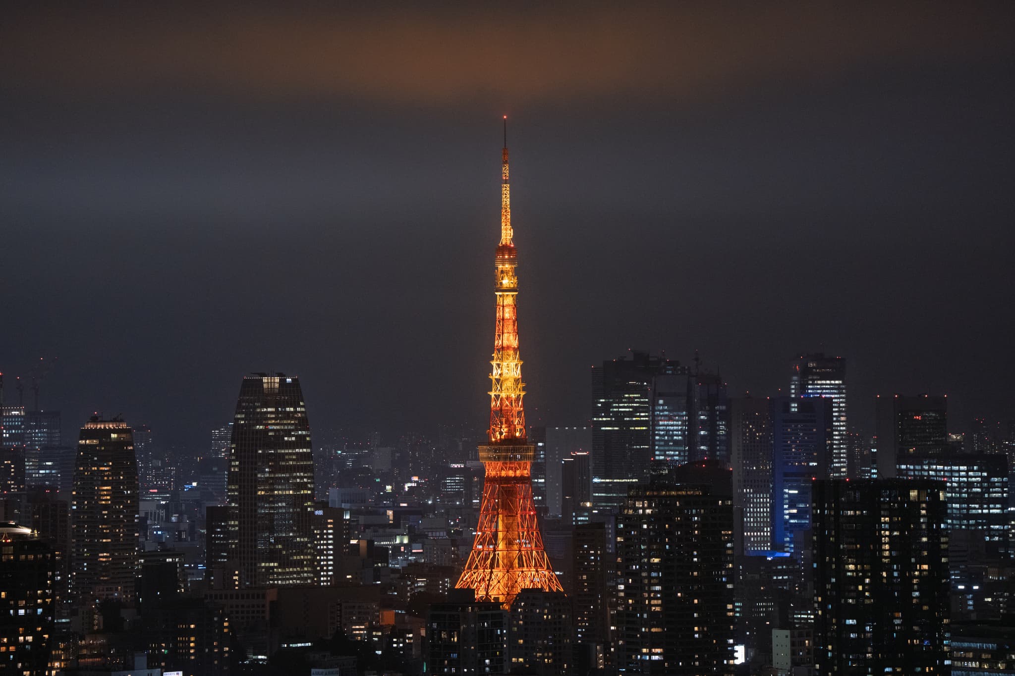 夜晚的城市天际线中，一座高塔被橙色灯光照亮，周围环绕着高楼大厦，天空阴沉。