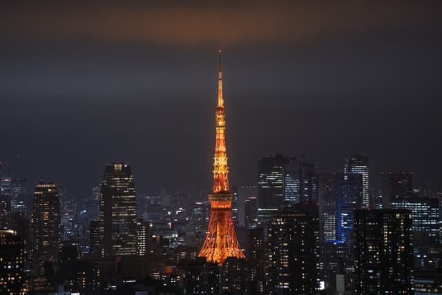 夜晚的城市天际线中，一座高塔被橙色灯光照亮，周围环绕着高楼大厦，天空阴沉。